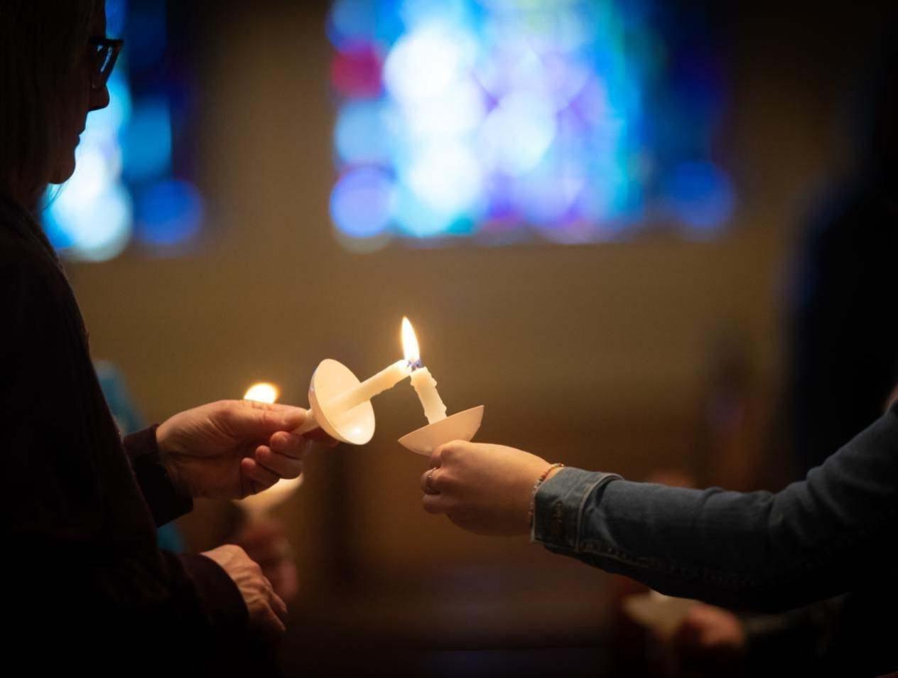 In a dimly lit room, one person uses their taper candle to light another person's candle.